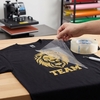 Hand die de transparante POLI-TACK 854 applicatiefolie verwijdert van een zwart t-shirt bedrukt met gouden leeuwenmotief